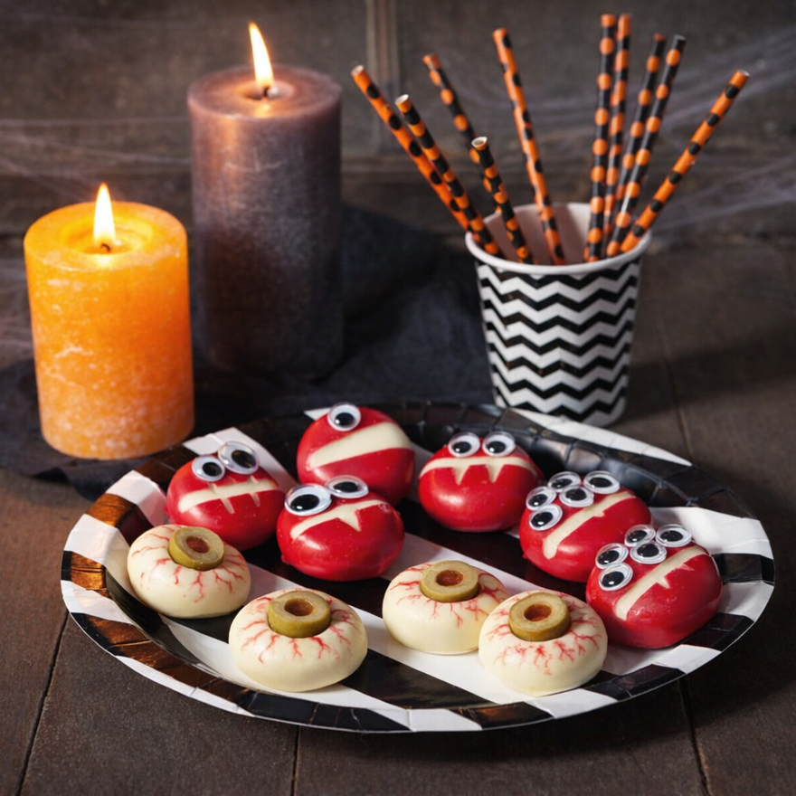 Rote und weiße Babybel-Käse mit Zahnreihen, Wackelaugen und Oliven-Pupillen auf einem gestreiften Teller neben brennenden Kerzen.