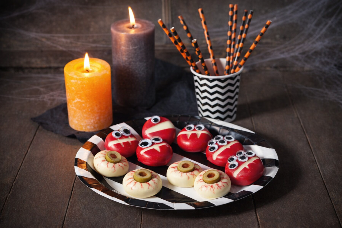 Rote und weiße Babybel-Käse mit Zahnreihen, Wackelaugen und Oliven-Pupillen auf einem gestreiften Teller neben brennenden Kerzen.
