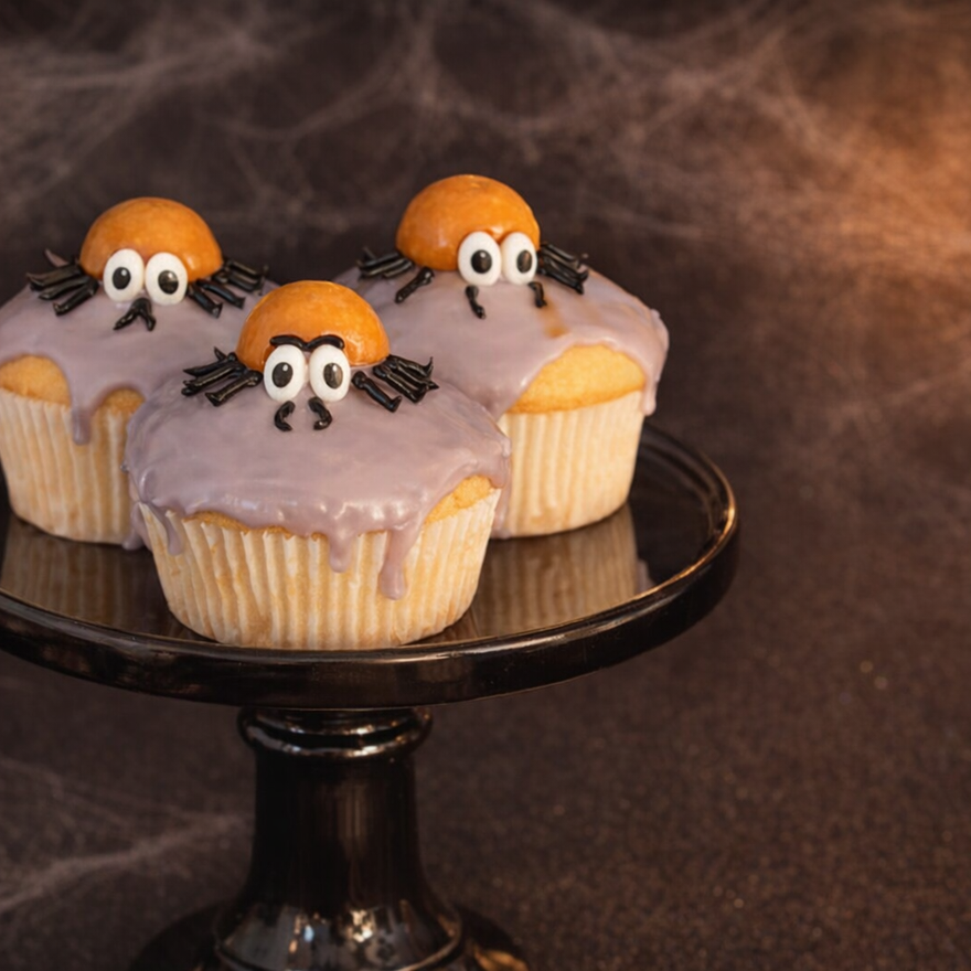 Drei Muffins mit lila Zuckerguss, orangefarbenem Kürbiskopf, Wackelaugen und schwarzen Lakritz-Beinen als Spinnen auf einem schwarzen Tortenständer.